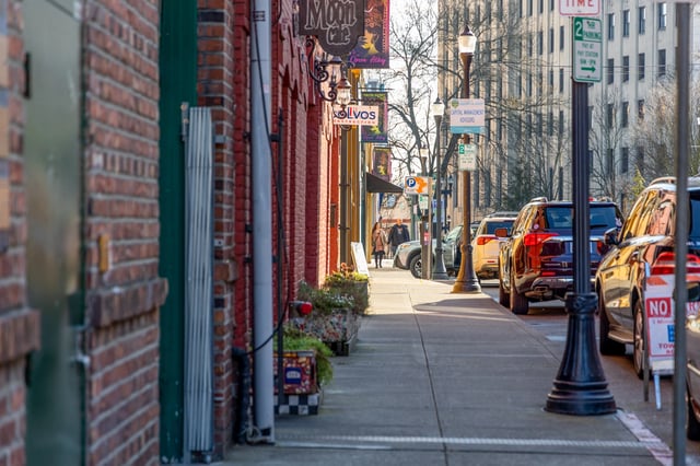 Street view of a brick building facade with shops and parked cars along the sidewalk.