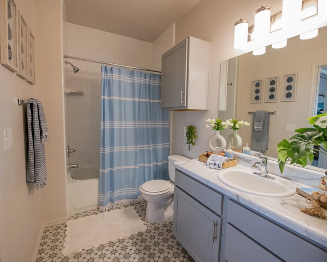 Bathroom in an apartment with marble vanity, light blue cabinetry, blue striped shower curtain, and geometric floor tiles.