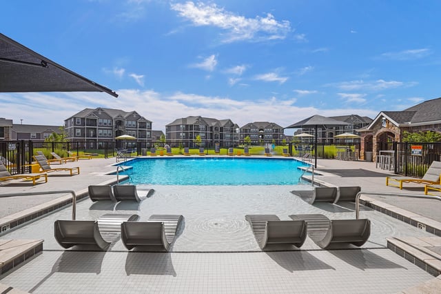 Resort-style swimming pool with in-water loungers and lounge chairs, surrounded by apartment buildings.