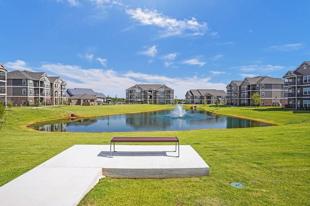 Apartment complex with a pond and fountain, surrounded by manicured lawns and trees.