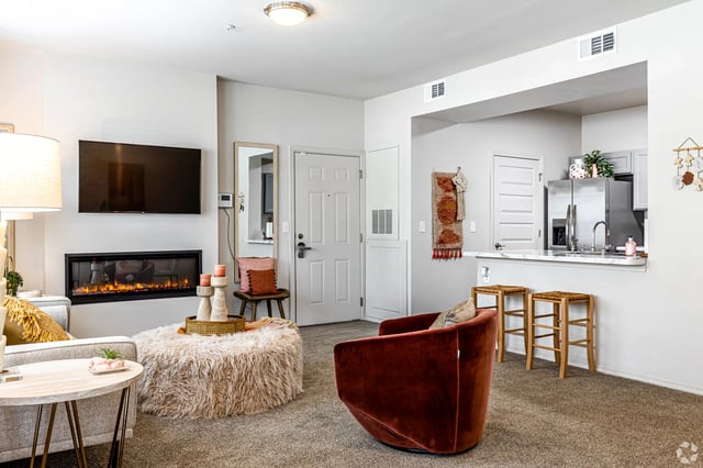 Modern living room with fireplace, TV, plush ottoman, and open kitchen view with bar stools.