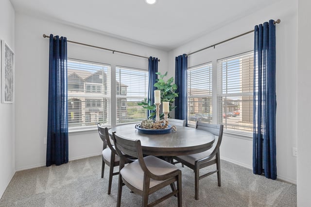 Dining room with a round table and four chairs, featuring blue curtains and a view of the exterior buildings.