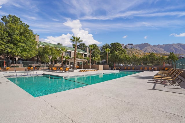 Outdoor swimming pool with lounge chairs and palm trees, mountains in the background.