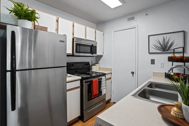 Stainless steel refrigerator and oven in a brightly lit kitchen with white cabinets and a large sink.