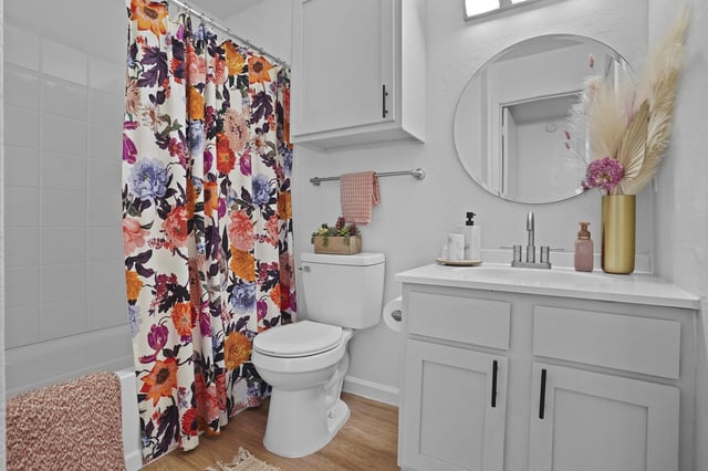 Bathroom with white vanity, round mirror, colorful floral shower curtain, toilet, and wood-look flooring.