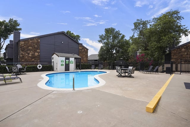 Outdoor apartment community pool with lounge chairs, tables, and a surrounding fence.