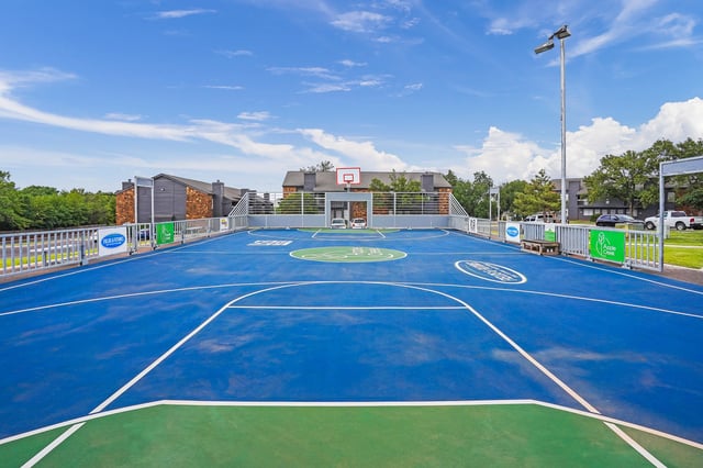 Bright blue basketball court with green accents, fencing, and apartment buildings in the background.