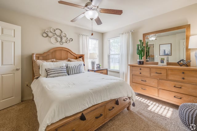 Bedroom with wooden bed, matching dresser, large mirror, ceiling fan, and two windows with white curtains.