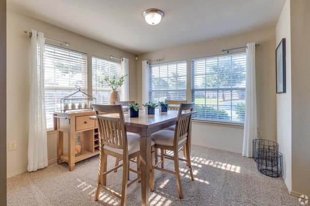 Dining area with a wooden table and four chairs beside large windows with sheer curtains.
