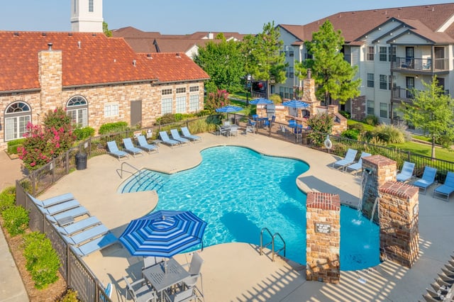 Outdoor community pool area with lounge chairs, umbrellas, and brick seating features beside apartment buildings.