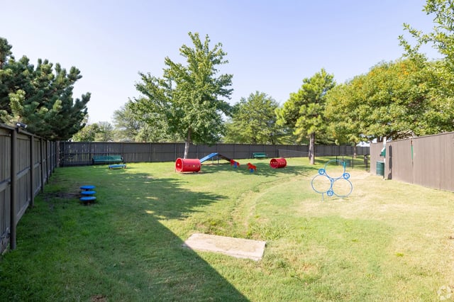 Fenced grassy community playground with red tunnels, a slide, and other play equipment.