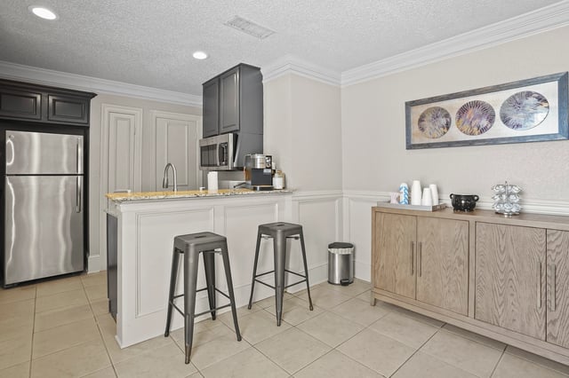Community coffee and refreshment station with a stainless steel refrigerator, microwave, coffee maker, and a counter with stools.