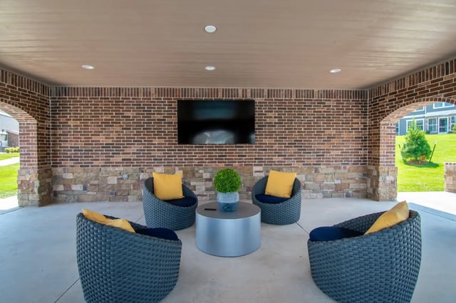 Outdoor covered lounge with brick walls, four blue wicker chairs with yellow cushions, and a mounted TV.