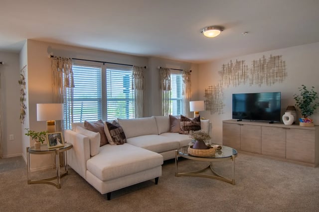 Living room with a white sectional sofa, glass coffee table, TV, and large windows with blinds.