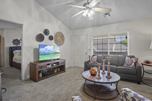 Living room in an apartment featuring a gray sofa, round coffee table, TV stand, ceiling fan, and window.