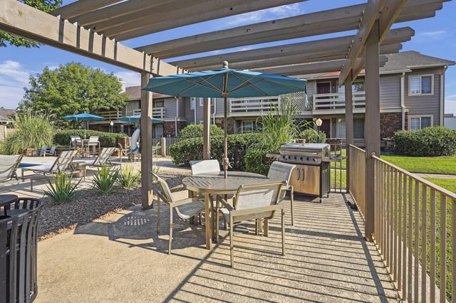 Outdoor communal patio with table and chairs, umbrellas, a grill, and lounge seating near buildings.