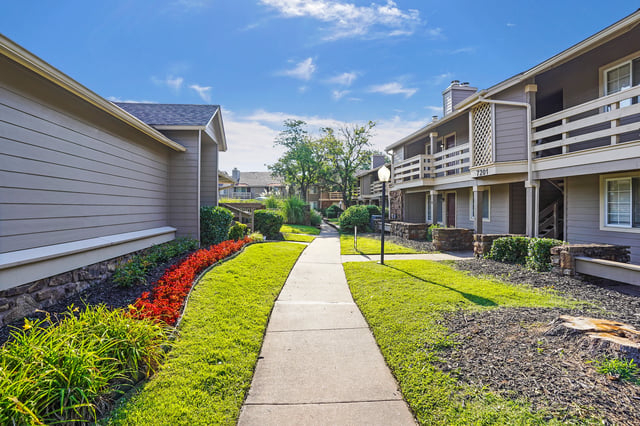 A paved walkway leads through a grassy courtyard with manicured flower beds between apartment buildings.