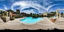 Panoramic view of a swimming pool area with lounge chairs and umbrellas, surrounded by buildings and landscaping.