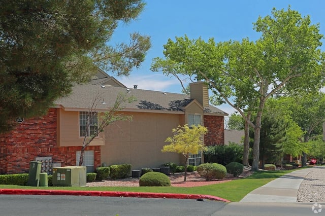 Exterior view of apartment buildings with brick and tan siding, surrounded by green trees and landscaping.