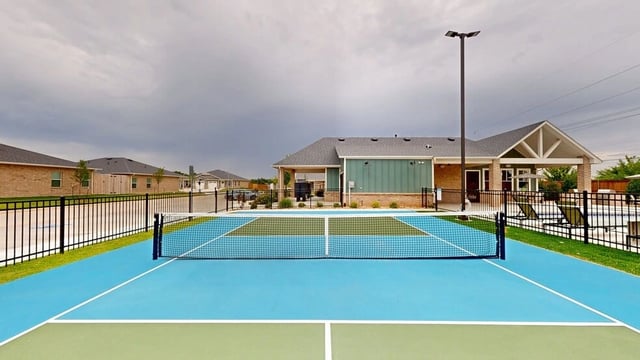 Outdoor tennis or pickleball court with a net, fenced in with apartment buildings in the background.