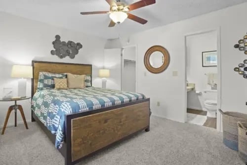 Queen bed with blue and white patterned comforter, flanked by bedside tables. A doorway reveals a bathroom with a toilet and vanity.