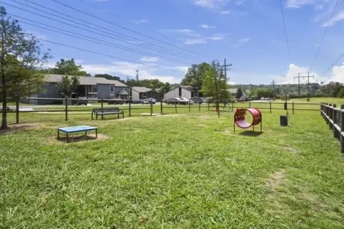 Open grassy community park with a playground tunnel, benches, and fencing near apartment buildings.