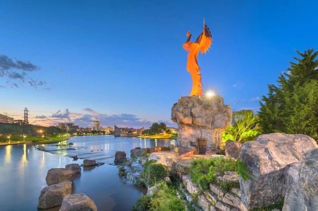 Evening view of a landscaped lakeside park with a tall orange statue on a rock pedestal.