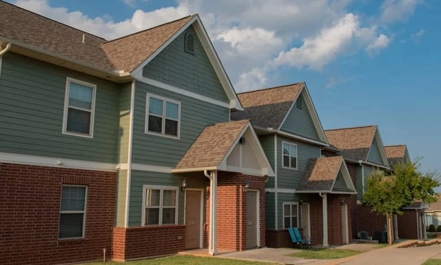 Exterior view of green siding apartment buildings with brick lower façade and entry doors along a sidewalk.