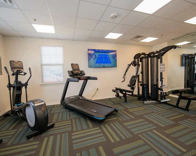 Interior of a community gym with treadmills, ellipticals, weight machines and a wall-mounted TV.