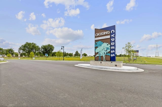 Entrance sign for Crossing Cypress Luxury Apartments at a roundabout, with landscaping and blue sky.