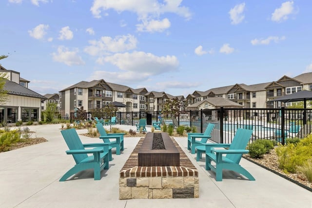 Outdoor communal pool area with a brick fire pit and turquoise Adirondack chairs, surrounded by apartment buildings.