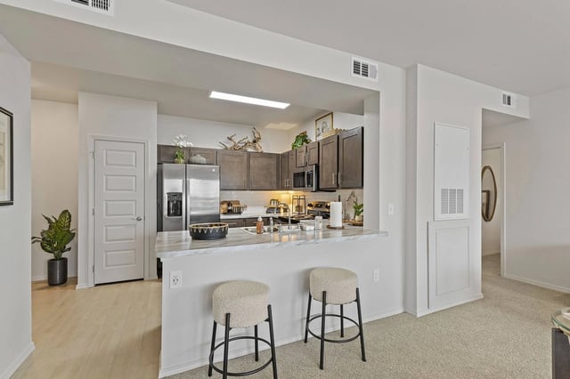 Open-concept kitchen with marble island, dark cabinets, stainless steel appliances, and two beige stools.