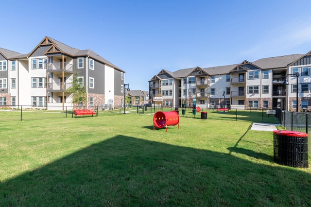 Apartment buildings with a fenced-in dog park area in the foreground, featuring a red tunnel and benches.