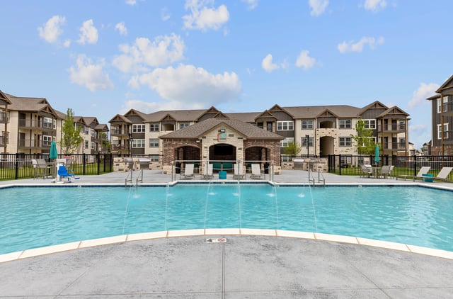 Outdoor swimming pool with fountains and lounge chairs, with apartment buildings in the background.