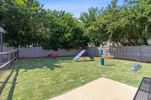 Fenced community playground with a slide, seesaw, and climbing structure on a grassy lawn.
