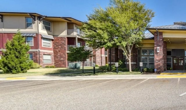 Apartment building exterior with brick and red accents, balconies, and manicured trees.