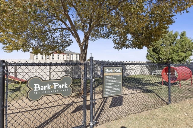 Fenced dog park at an apartment community with a large tree, signs, and a red tunnel.