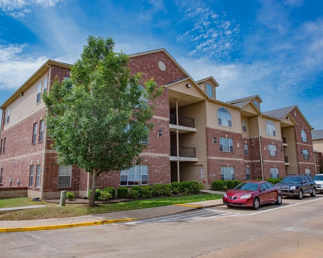 Exterior view of a brick-and-beige apartment building with balconies, parking, and landscaping.