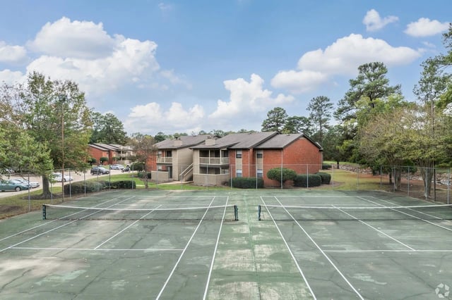 Outdoor tennis court with white lines and brick apartment buildings in the background.