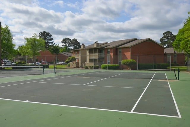 Outdoor tennis court with a net, adjacent to brick apartment buildings and manicured landscaping.