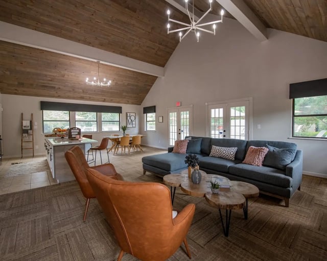 Open-concept community lounge with sofa, chairs, kitchen island, and dining table under wood-beamed ceiling.