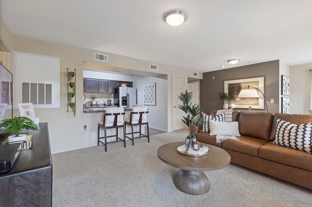 Open-concept living area with beige walls, carpet, a brown leather sofa, and a kitchen bar in the background.