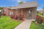Exterior of a small building with red brick and a porch, surrounded by green grass and red flowers.