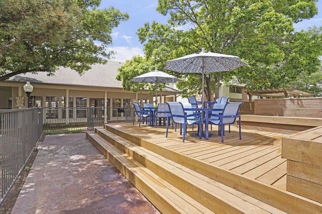 Outdoor community deck with blue chairs, umbrellas, steps, and railing under trees.