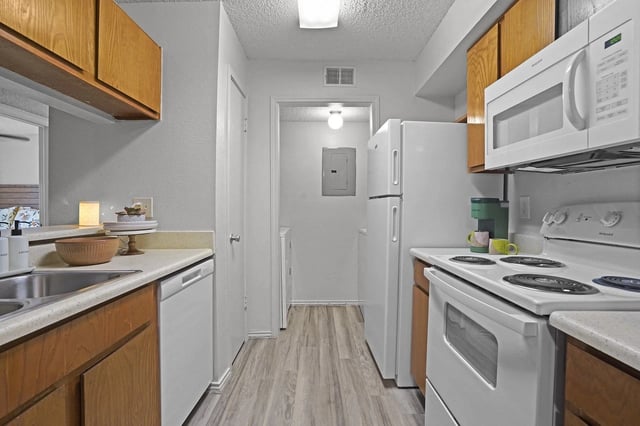 Galley-style apartment kitchen with white appliances, wooden cabinets, and light vinyl flooring.
