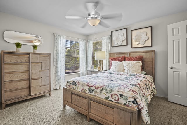 Bedroom with wooden bed frame, matching dresser, windows with white curtains, and a ceiling fan.