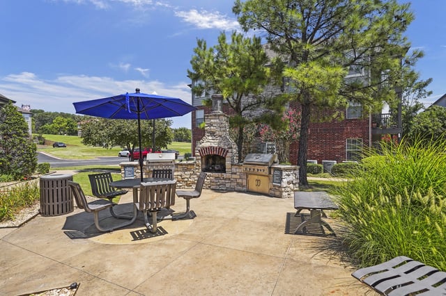 Outdoor resident gathering space with a stone grill, table and bench seating, umbrella, and landscaping near the apartment building.
