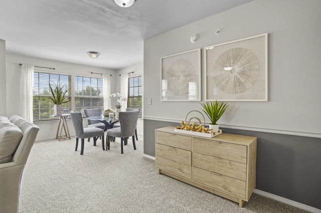 Bright living/dining area with large windows, a glass round table, four grey upholstered chairs, and a wood sideboard.