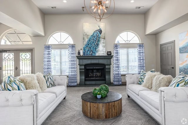 Community lounge with white tufted sofas, a dark fireplace, peacock artwork, and arched windows.
