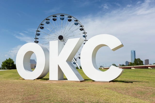 Large white OKC monument letters on a grassy field with a ferris wheel and city skyline.